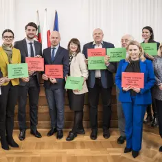 Grupa osób w garniturach i strojach biznesowych trzyma tabliczki z nazwami projektów miejskich.