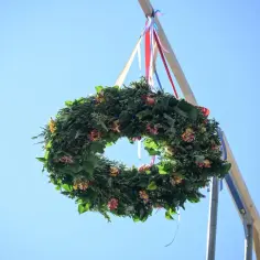 Wieniec z zieleni i kwiatów zawieszony na drewnianym słupie z kolorowymi wstążkami na tle błękitnego nieba.