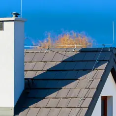 Biały komin i ciemny dach z dachówkami. Na dachu widoczna jest odgromówka, chroniąca dom przed burzą.
