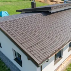 Nowy dom z brązowym, falistym dachem z miedzi. W oddali widać niebieski namiot na zielonym trawniku.