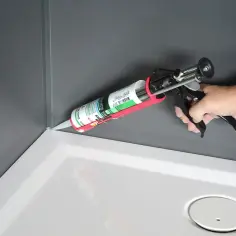 Dłoń z pistoletem wyciska dobry silikon do kabiny prysznicowej, uszczelniając narożnik brodzika.