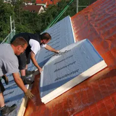 Dwóch budowlańców układa płyty izolacyjne na dachu. To kluczowy etap izolacji dachu od zewnątrz, zapewniający komfort termiczny.