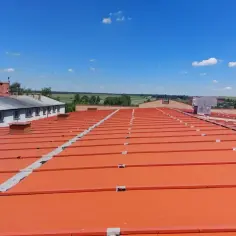 Pomarańczowy, lekki dach płaski z metalowymi elementami, rozciągający się pod błękitnym niebem z chmurami.