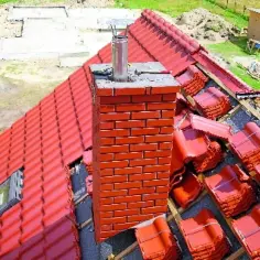 Trwa montaż dachówki, a komin czeka na obróbki komina blachą. Widać stosy dachówek i fundamenty nowego domu.