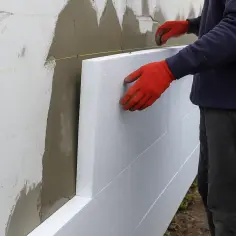 Ręce w czerwonych rękawiczkach przyklejają biały styropian do ściany. Zastanawiasz się, ile kosztuje przyklejenie styropianu?