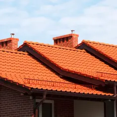 Pomarańczowy dach z dachówki ceramicznej, z ceglanymi kominami i brązowymi rynnami, pod błękitnym niebem.