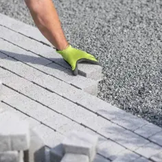 Ręka w zielonej rękawicy układa kostkę brukową na podsypce. To kluczowy etap, by wybrać odpowiedni cement pod kostkę.