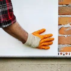 Ręka w pomarańczowej rękawicy przykłada białe płyty PIR do ceglanej ściany. To izolacja poddasza.