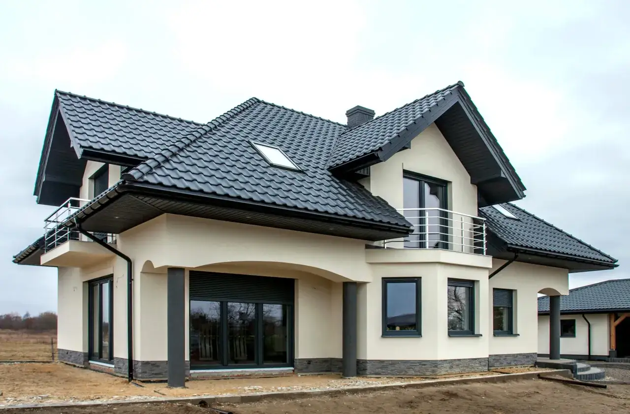 Nowy dom z jasnym pucem na elewacji i czarnym dachem. Widać balkon i okna z roletami.