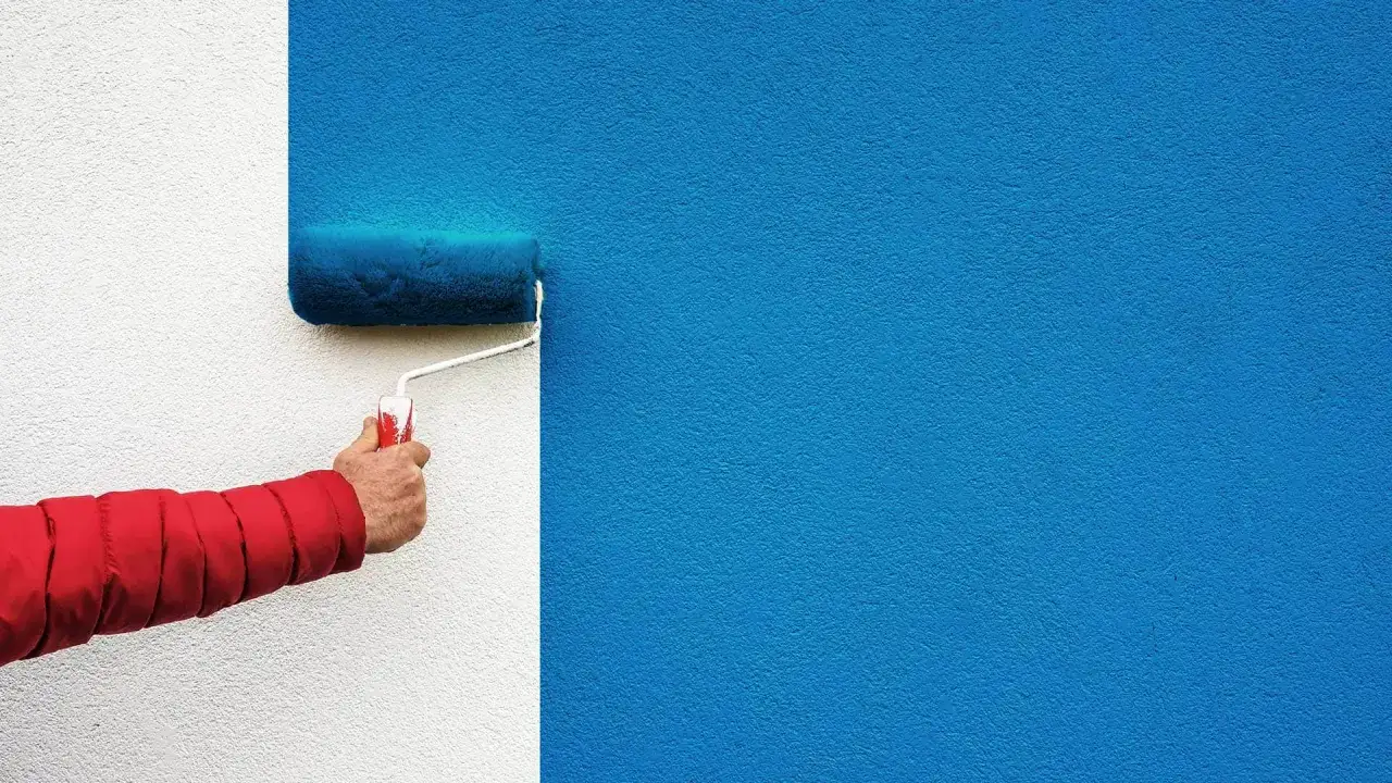 Ręka w czerwonej kurtce maluje ścianę niebieską farbą, używając wałka. Różne rodzaje wałków do malowania pozwalają uzyskać różne faktury.