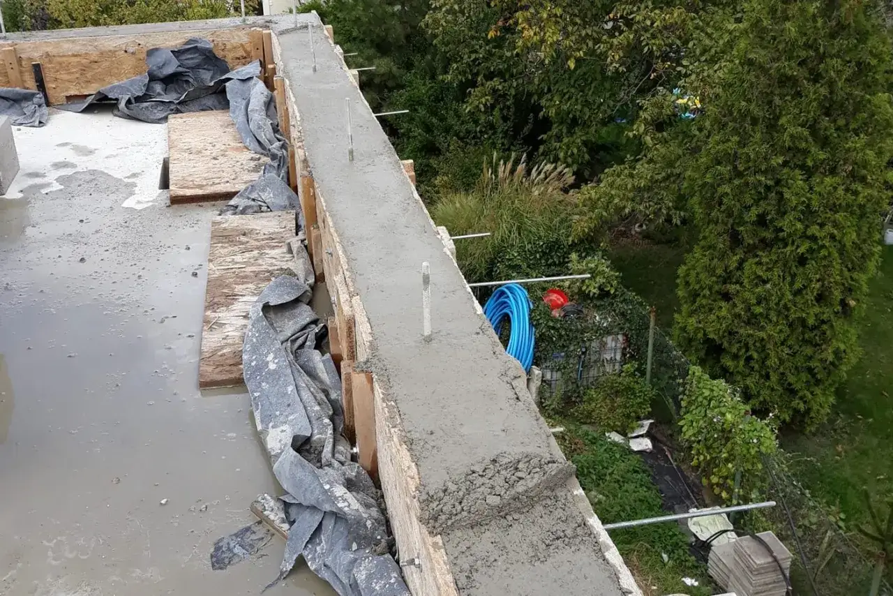 Świeży beton na stropie, widać szalunki i kilka szpilek do murlaty. W tle ogród z drzewami i niebieskim wężem.