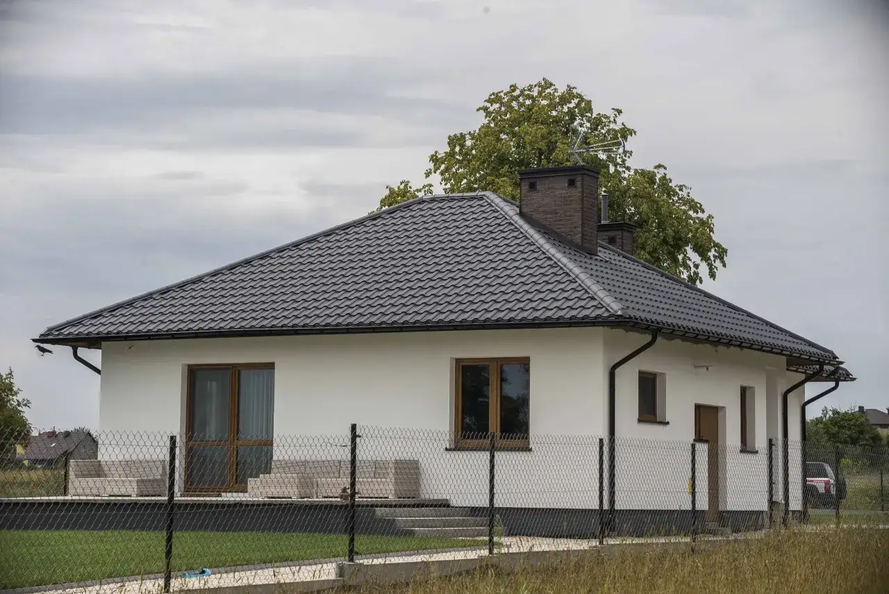 Nowy dom z białymi ścianami i ciemnym dachem z płyty obornickiej. Za płotem widać zielony trawnik i drzewa.