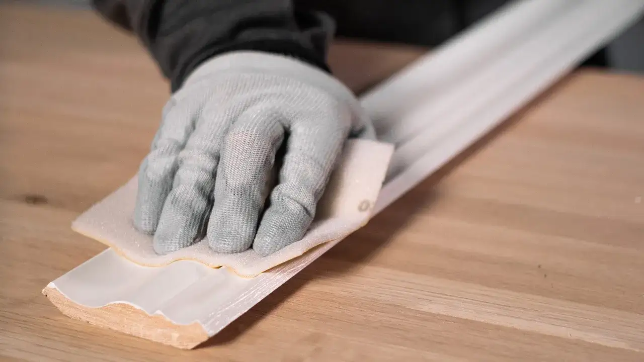 Ręka w rękawicy szlifuje białą listwę, używając papieru ściernego. Idealny papier do szlifowania drewna zapewnia gładkie wykończenie.
