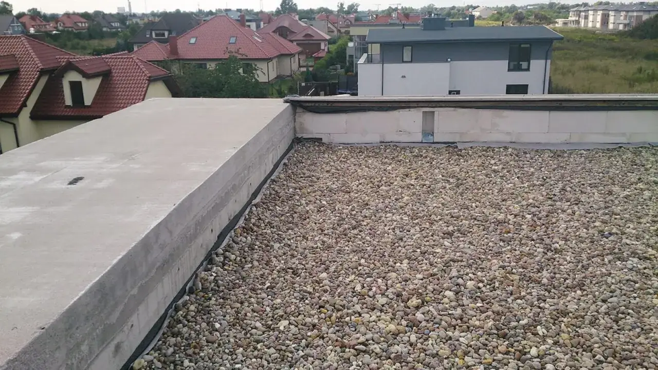 Na płaskim dachu leżą kamienie, tworząc warstwę ochronną. W tle widać domy z czerwonymi dachami.