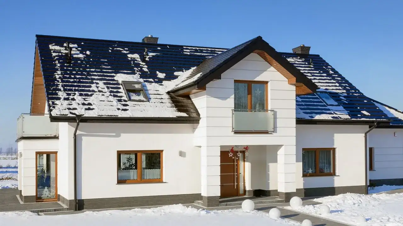 Nowy dom z białym tynkiem i czarnym, pokrytym śniegiem dachem z dachówki ceramicznej, otoczony zimowym krajobrazem.
