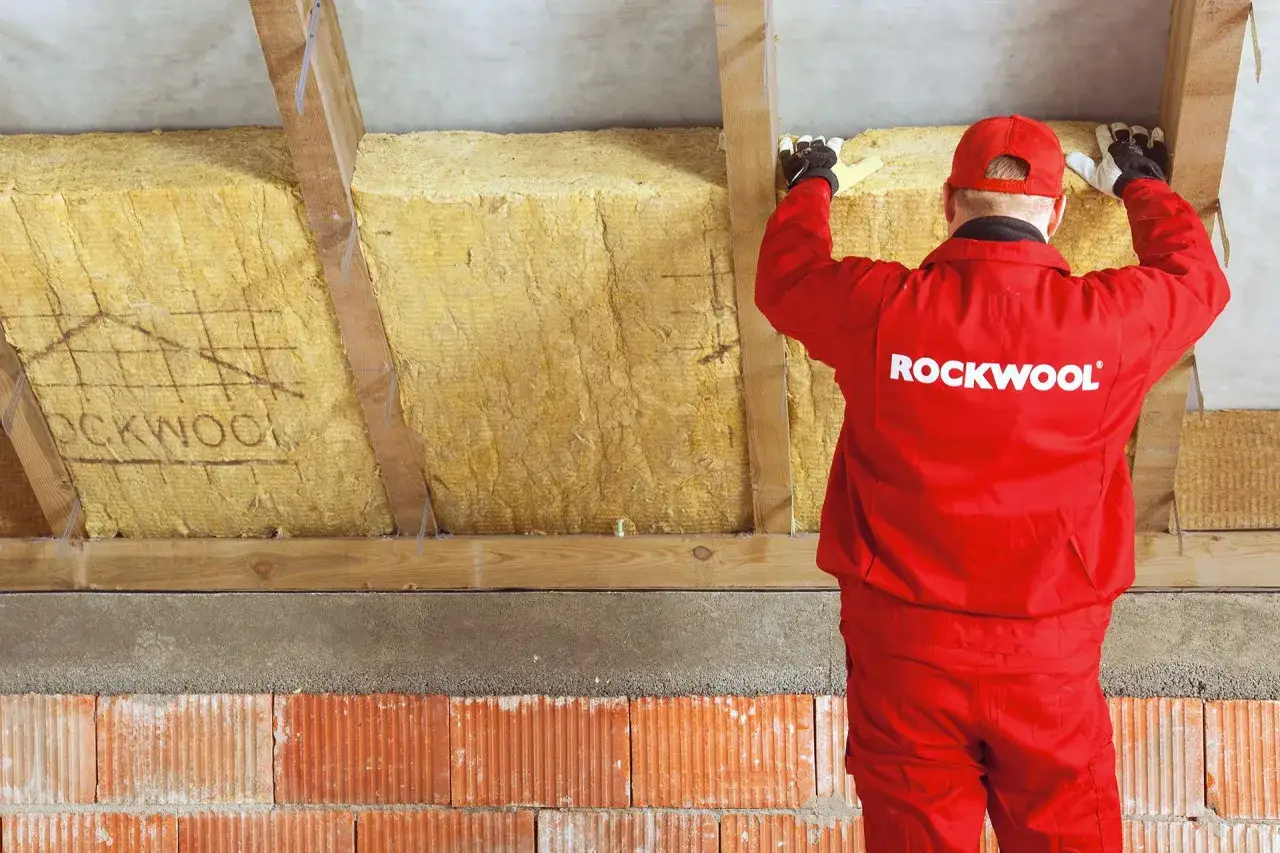 Pracownik w czerwonym kombinezonie Rockwool montuje wełnę mineralną w przestrzeni między murłatą a dachem.
