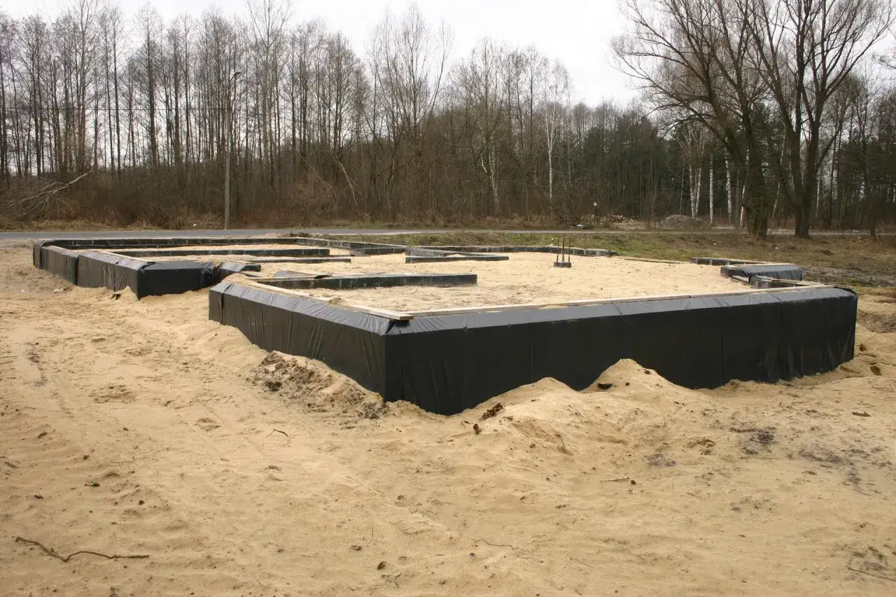 Czarne folie zabezpieczają fundament przed wilgocią, to kluczowe, czym zabezpieczyć fundament. Piasek otacza budowę.